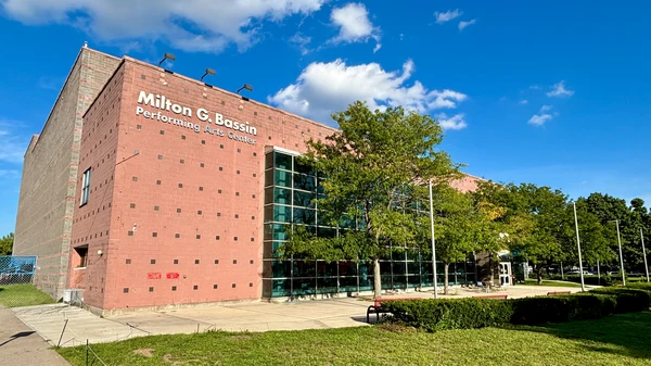 The exterior of the Milton G. Bassin Performing Arts Center, a large brick building with modern glass sections, under a bright blue sky.