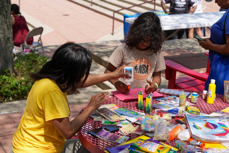 Adult and child work together on an arts-and-crafts activity at an outdoor table covered with colorful supplies, glue, markers, and paper projects.
