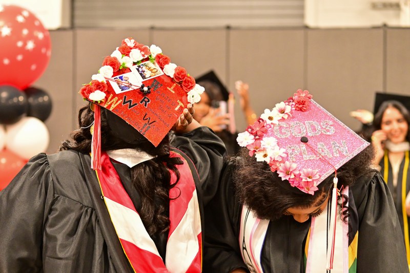 Two graduates in caps and gowns display decorated graduation caps adorned with flowers and personalized messages during a commencement celebration.