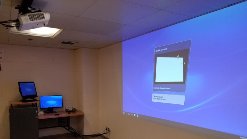 A picture of a class room with a projector, lectern, a projection screen and a computer.