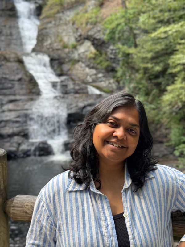Lecturer Shoba Parasram stands outdoors in front of a waterfall, discussing Writing Across the Curriculum.